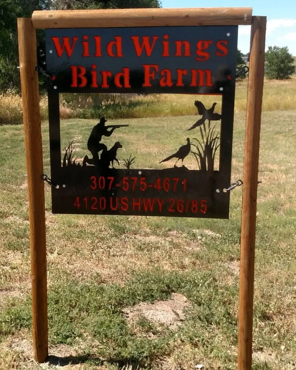 A metal sign for Wild Wings Bird Farm features silhouettes of a hunter, a dog, and pheasants.