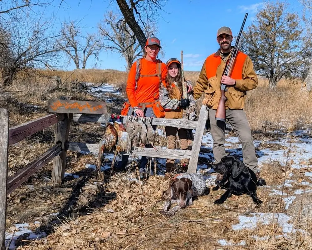 A family and two hunting dogs pose with harvested pheasants and chukar in a snowy field.
