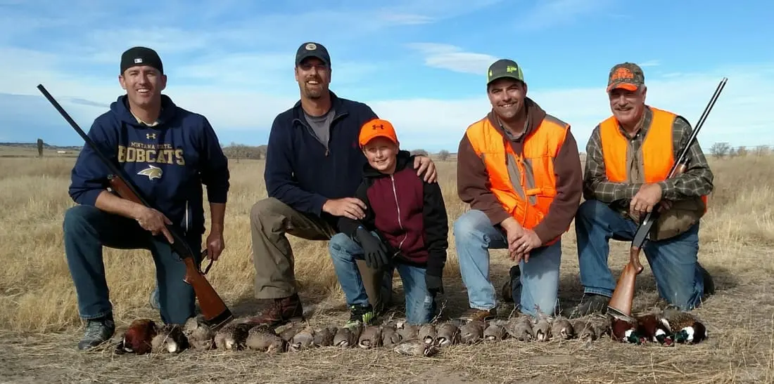 Five hunters, including one youth, pose with harvested pheasants and chukar in a field.