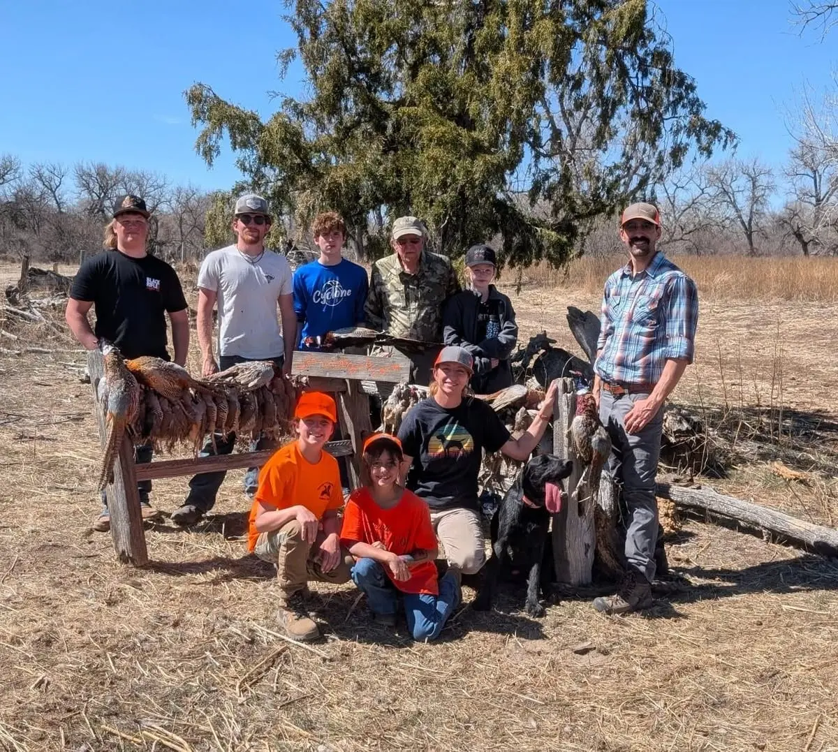A group of hunters, including adults and children, pose with their harvested pheasants and chukar in a field.