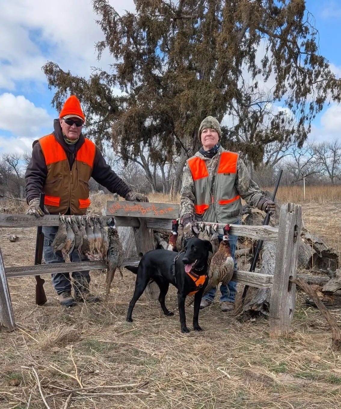 Two hunters and a black dog pose with harvested pheasants and chukar hanging on a wooden fence.