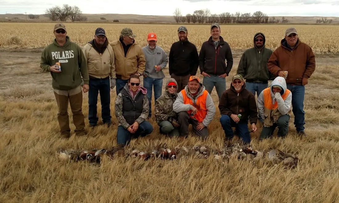 A group of hunters poses with their harvested pheasants and chukar in a field.