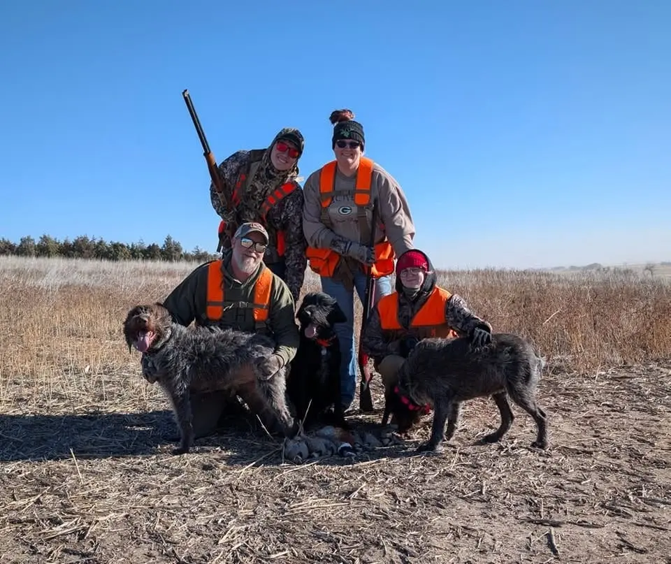 Four hunters and three dogs pose with harvested pheasants and chukar in a field.