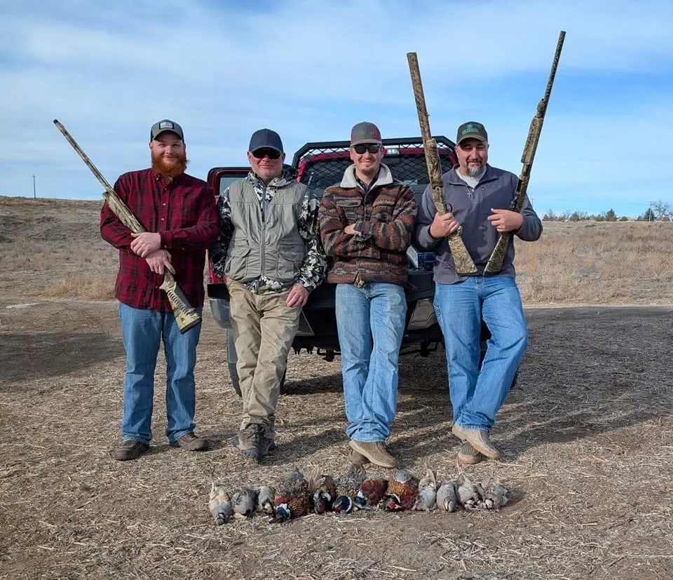 Four hunters pose with their harvested ring-necked pheasants and chukar partridges in a field.