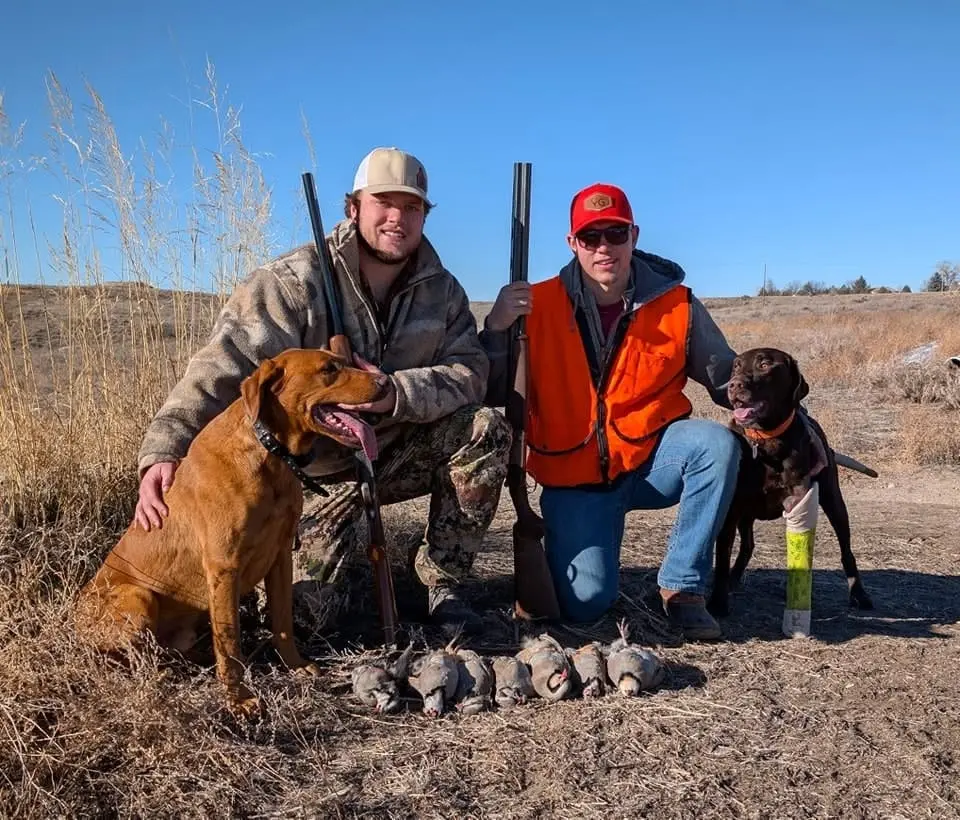 Two hunters pose with their dogs and several harvested chukar partridges in a field.