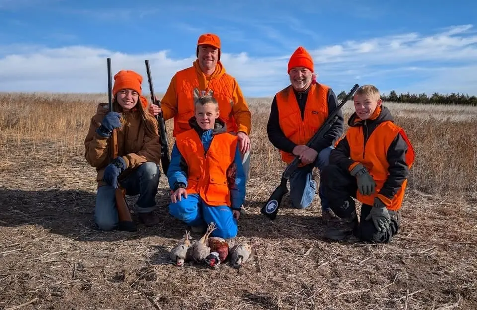 Five hunters, including adults and children, pose with harvested pheasants and chukar in a field.