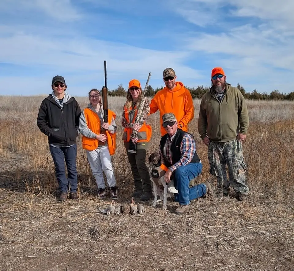 A group of hunters and a dog pose with harvested chukar in a field.