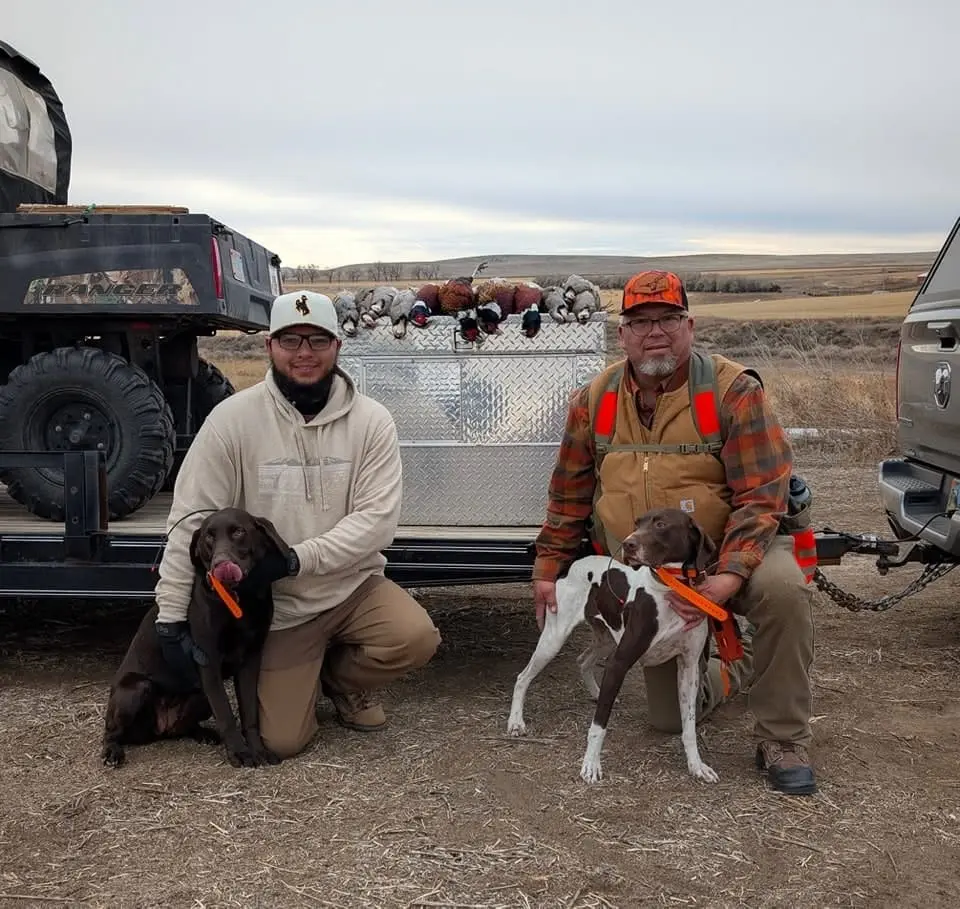 Two hunters pose with their dogs and a harvest of pheasants and chukar on a trailer.