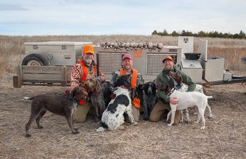Three hunters pose with their hunting dogs and harvested chukar in a field.