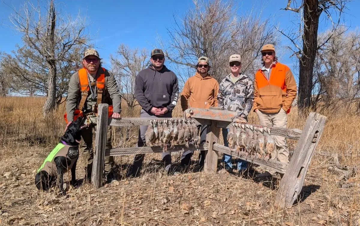 Five hunters and a dog pose with their harvest of chukar on a wooden rack in a field.