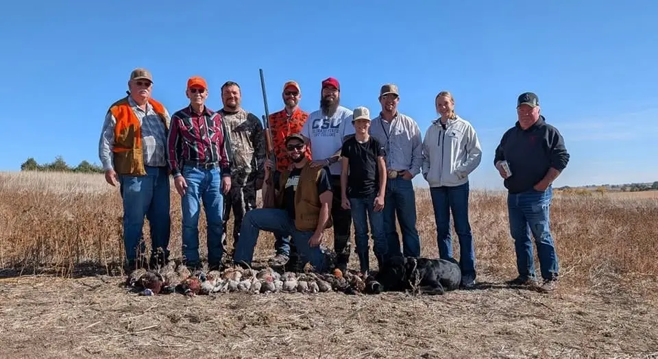 A group of hunters poses with their harvested pheasants and chukar in a field under a clear blue sky.