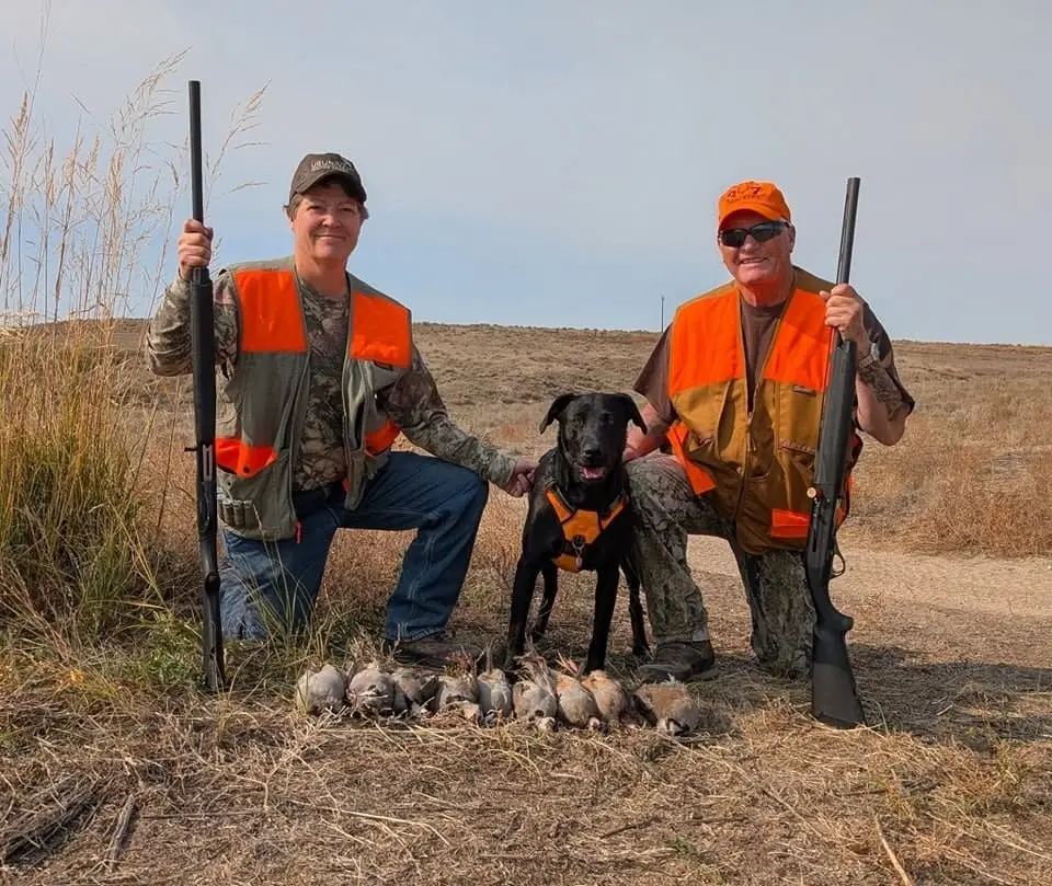 Two hunters and a dog pose with their harvested chukar partridges in a field.
