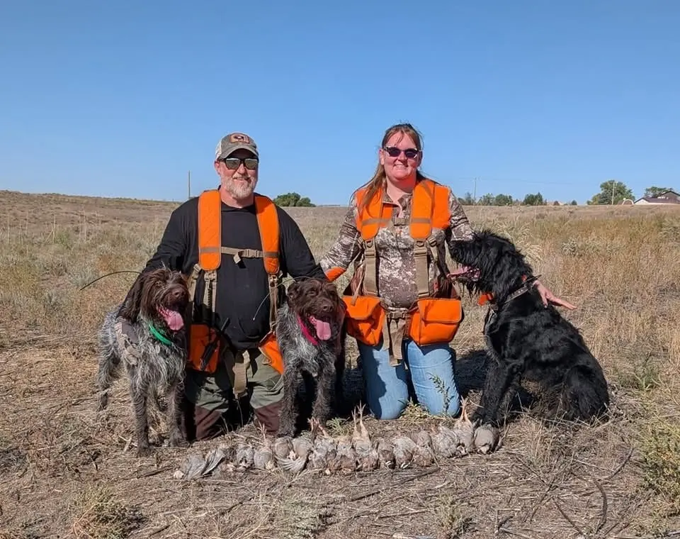 A man and a woman in hunting gear pose with three dogs and a pile of harvested birds in a field.
