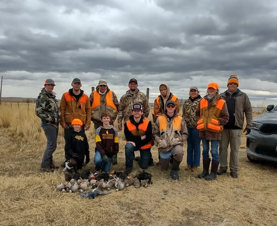 A group of hunters, including adults and children, pose with harvested ring-necked pheasants and chukar partridges on the ground.