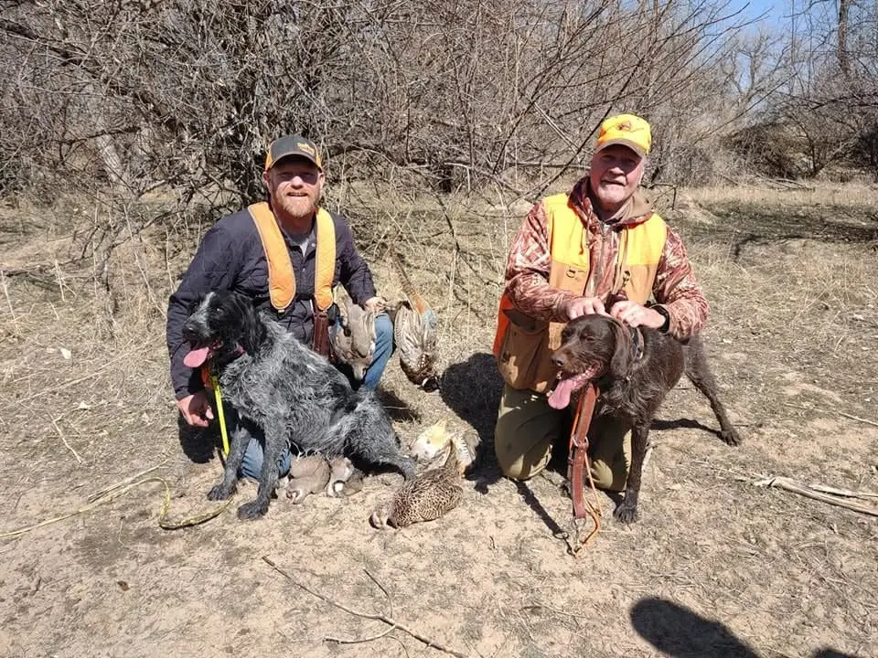 Two hunters pose with their dogs and several harvested pheasants and chukar in a field.