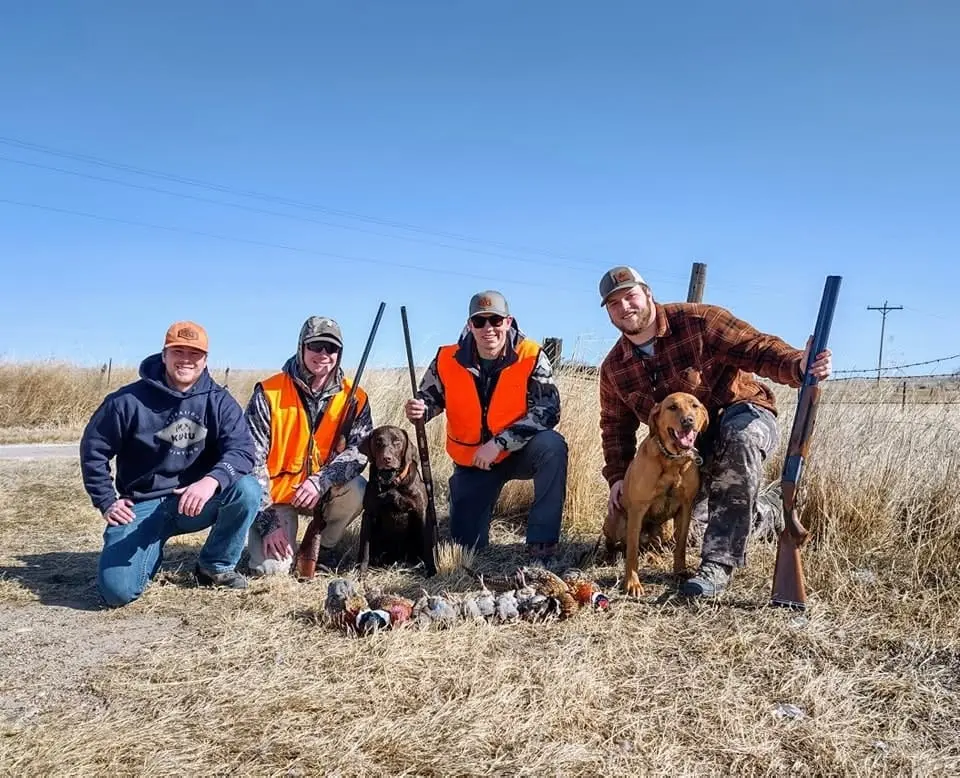 Four hunters and two dogs pose with harvested pheasants and chukar in a field.