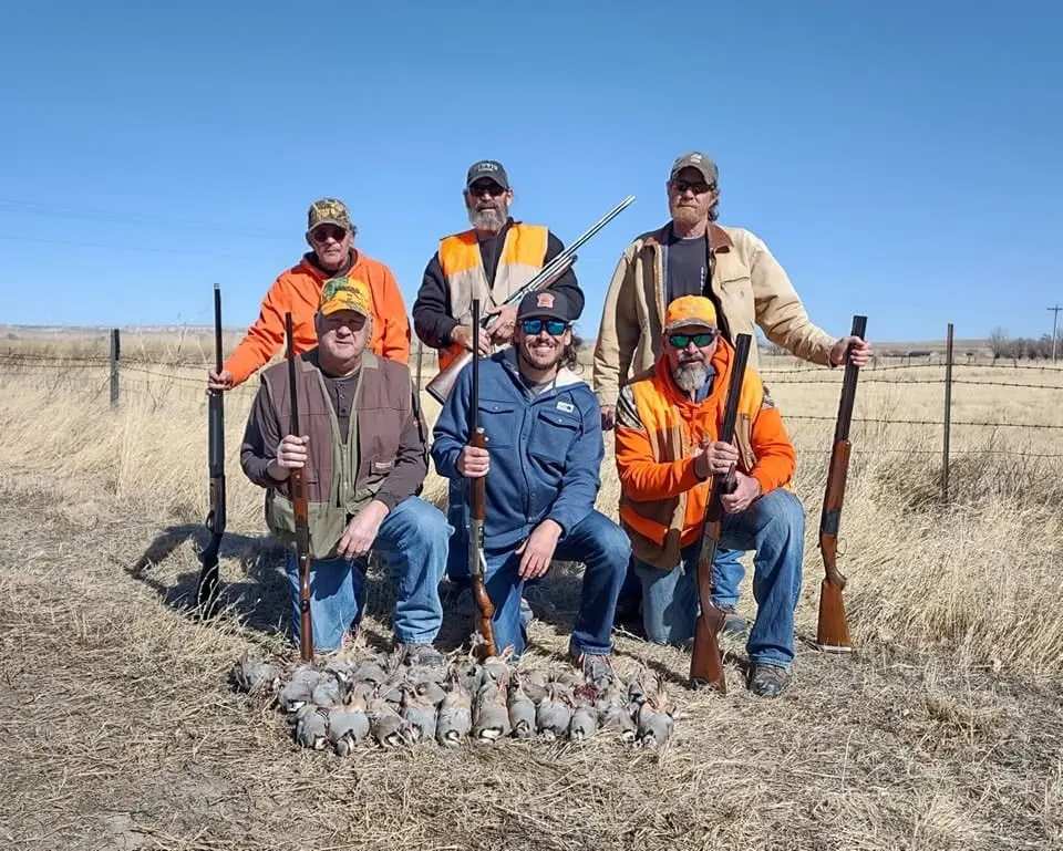 Six hunters pose with their shotguns and a pile of harvested chukar partridges in a field.