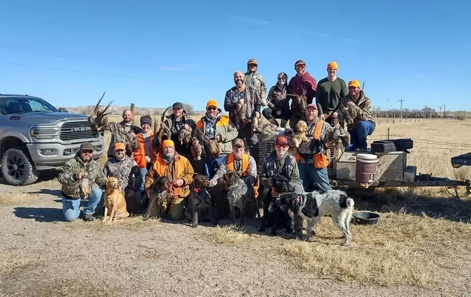 A large group of hunters and their dogs pose with harvested ring-necked pheasants next to a truck and trailer.