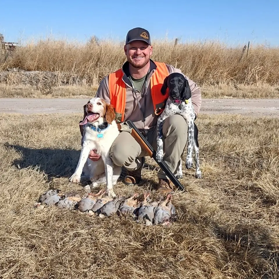 A hunter poses with two dogs and harvested birds in a field.