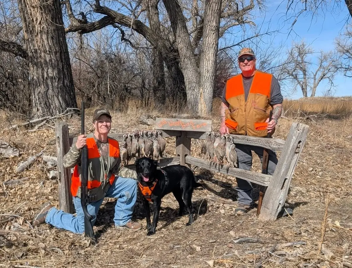 Two hunters and a dog pose with harvested chukar hanging on a wooden rack in a field.
