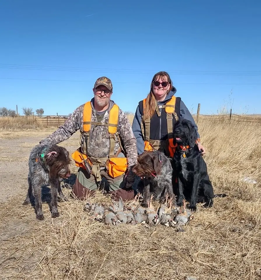 Two hunters pose with three dogs and a pile of harvested Chukar Partridges in a field.