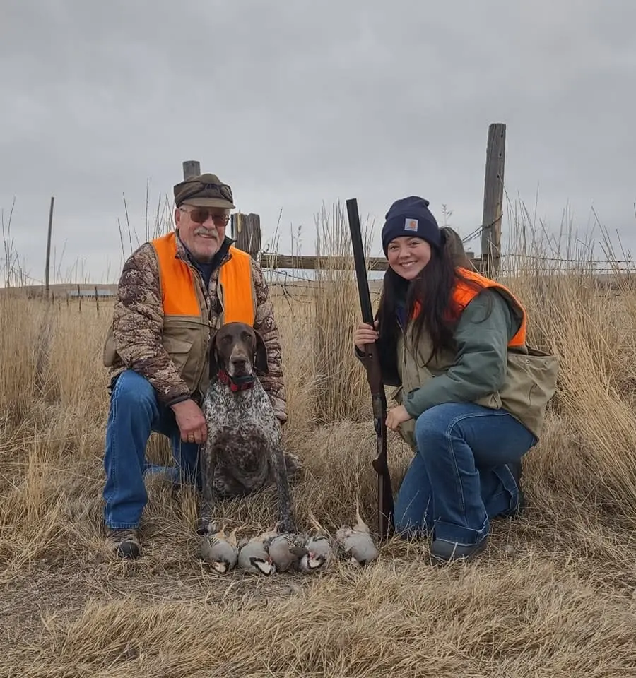 Two hunters and a dog pose with harvested chukar partridges in a field.