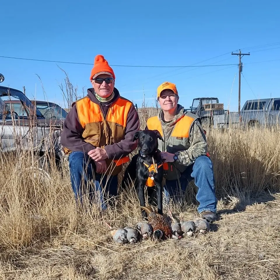 Two hunters and a dog pose with harvested ring-necked pheasants and chukar partridges in a field.