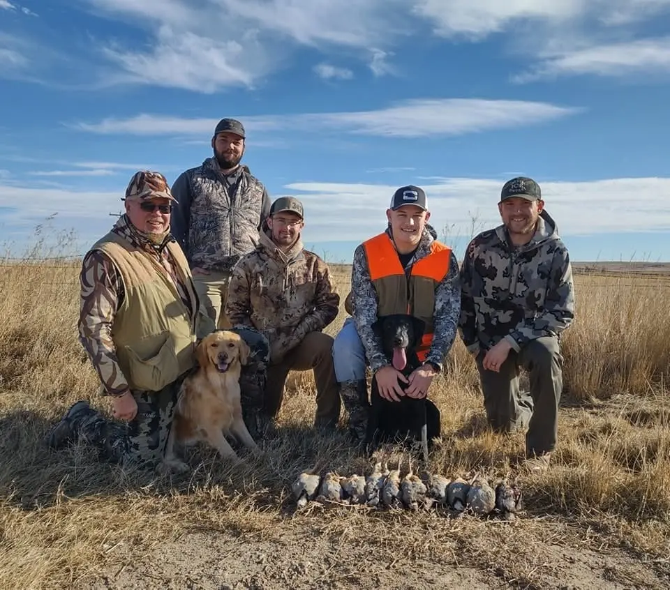 Several hunters pose with their dogs and a harvest of chukar in a field.