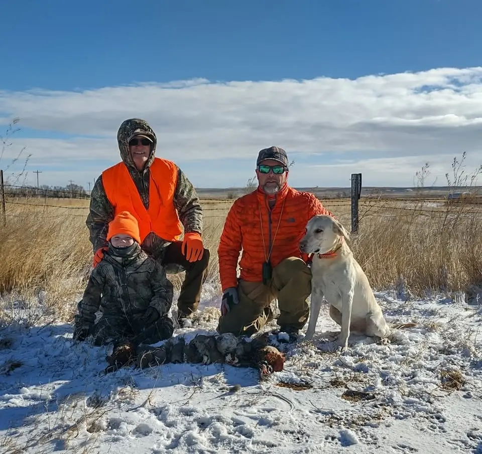 Three hunters and a dog pose with harvested pheasants and chukar in a snowy field.