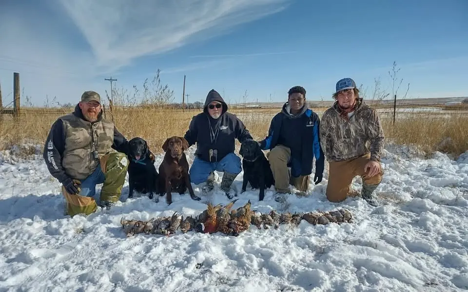 Four hunters and three dogs pose with their harvested pheasants in a snowy field.