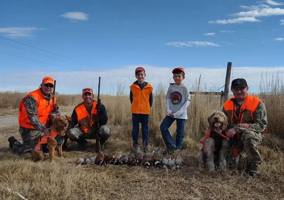 Hunters and two dogs pose with harvested ring-necked pheasants and chukar partridges in a field.