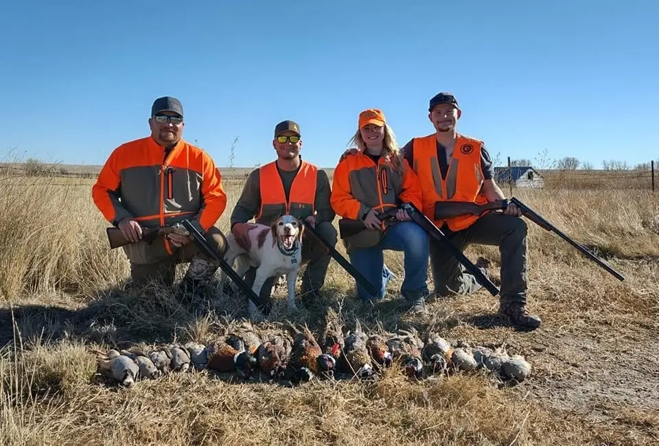 Four hunters and a dog pose with their harvest of pheasants and partridges in a field.