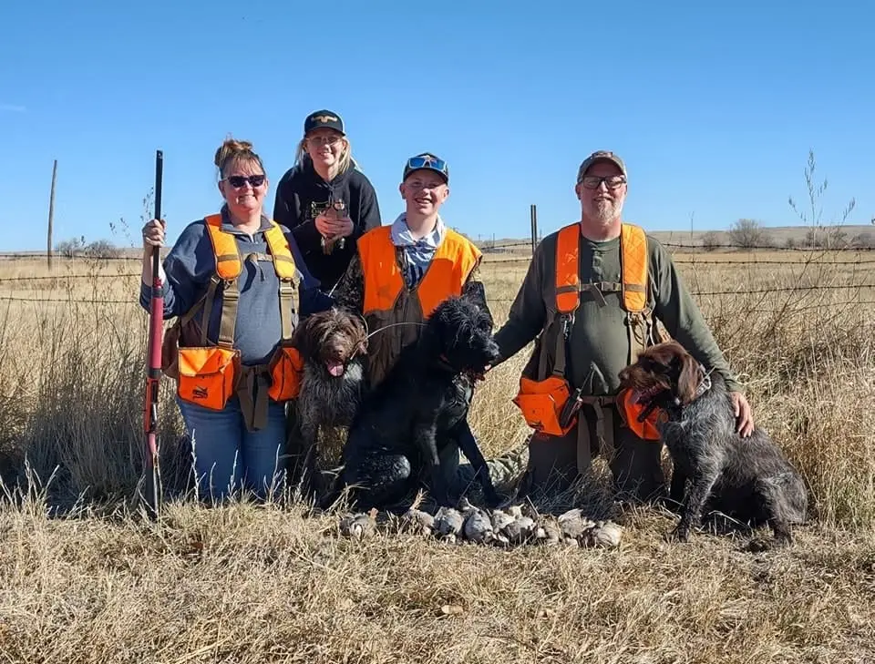 A group of hunters and their dogs pose with harvested chukar in a field.