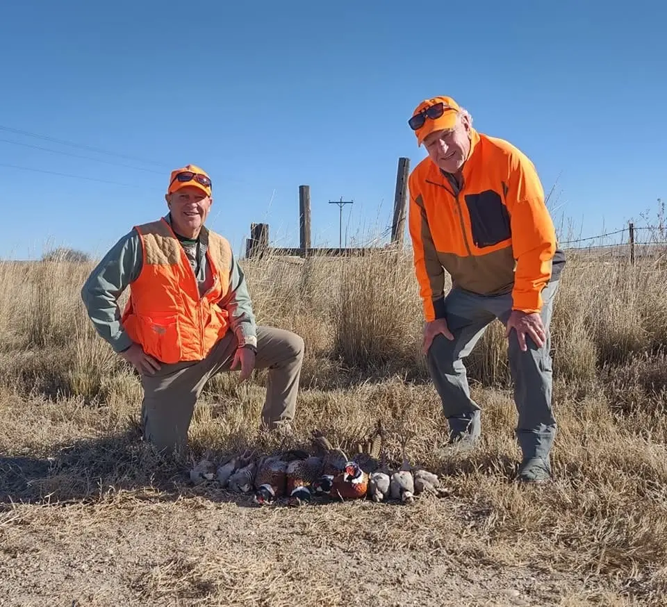 Two hunters pose with their harvested pheasants and chukar in a field.