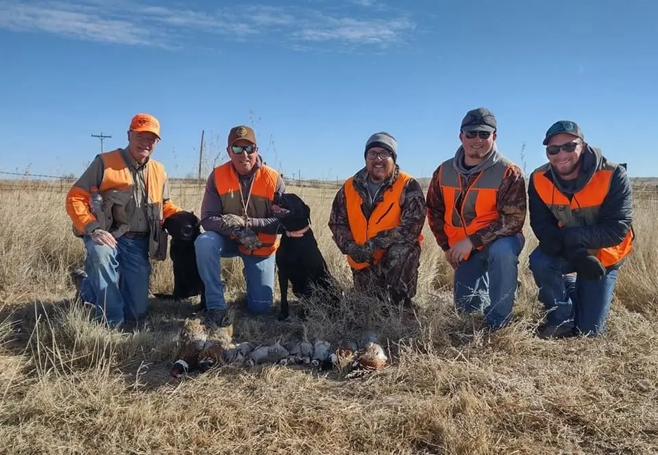 Five hunters and two dogs pose with harvested pheasants and chukar in a field.