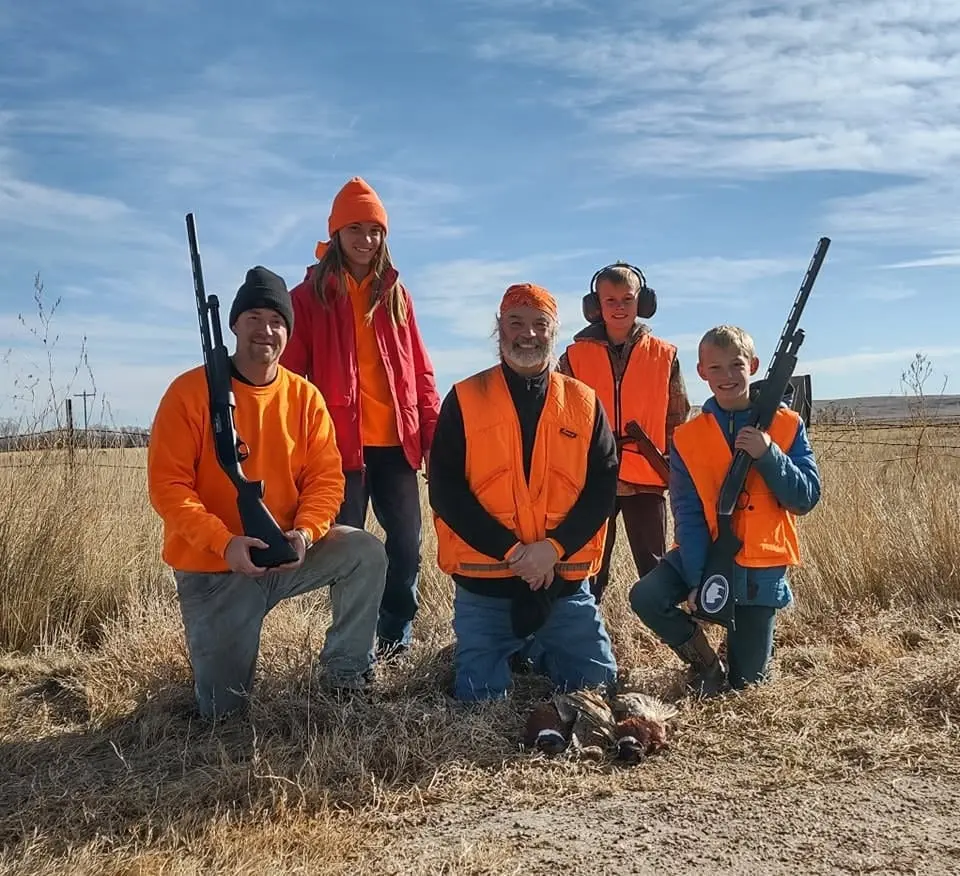 A group of five hunters, including children, pose with two harvested pheasants in a field.