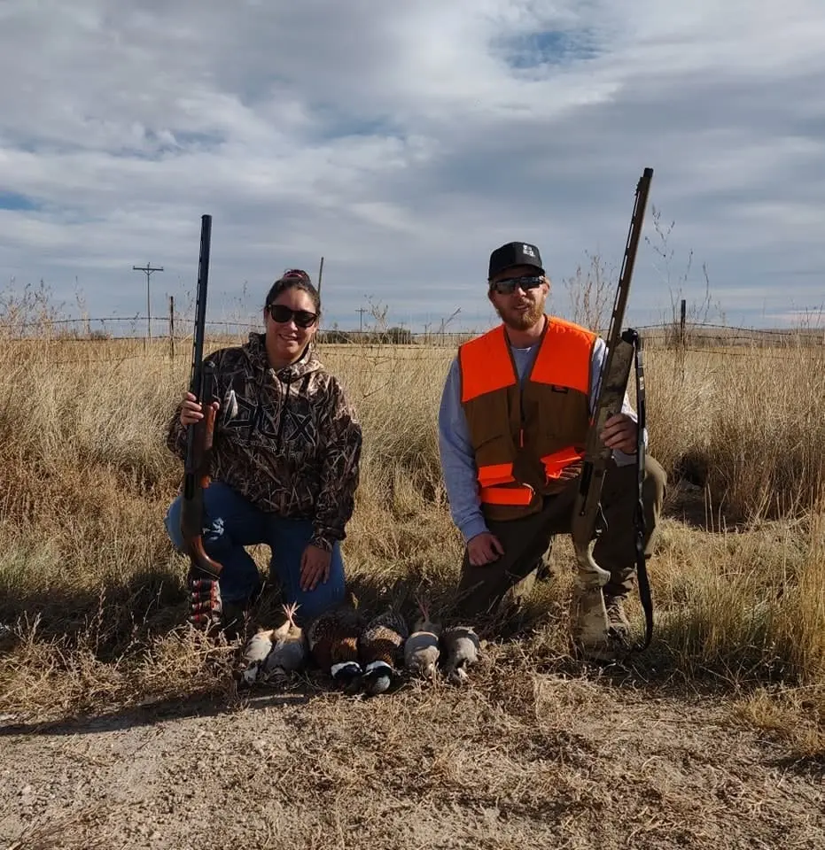 Two hunters pose with their harvested pheasants and chukar in a field.