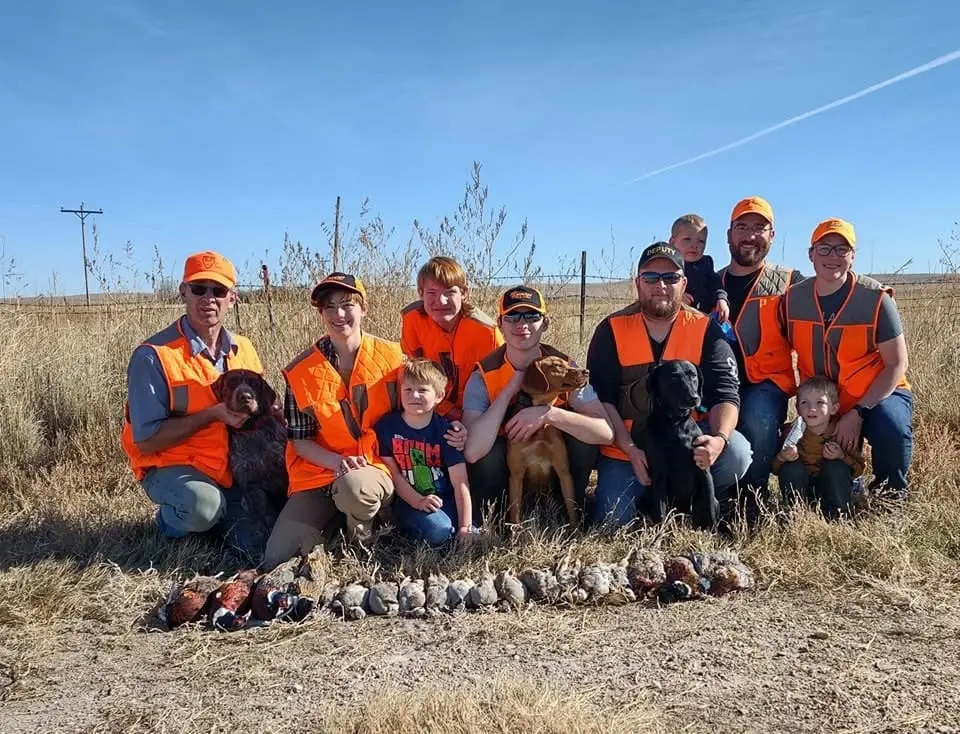 A group of hunters, including adults and children, pose with their hunting dogs and a harvest of ring-necked pheasants and chukar partridges in a field.