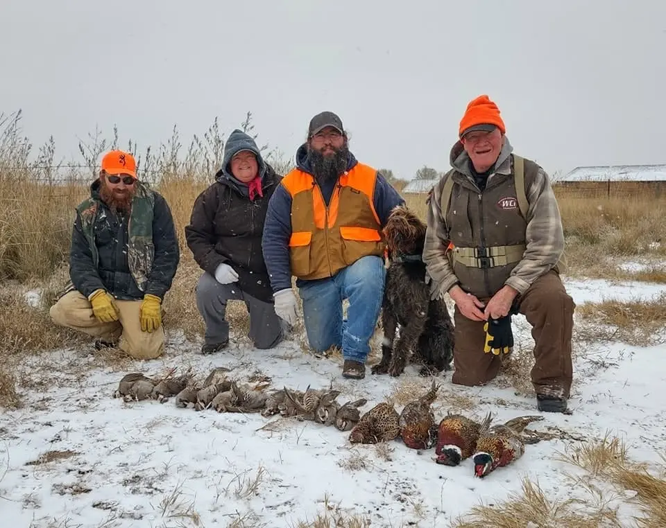 Four hunters and a dog pose with harvested pheasants and partridges in a snowy field.