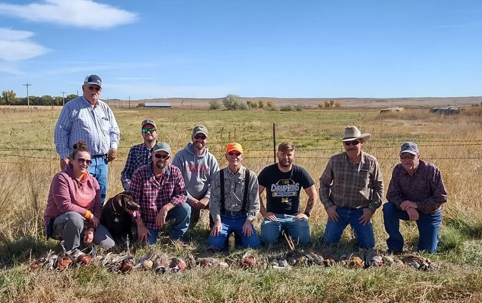A group of hunters and a dog pose with harvested ring-necked pheasants in a field.
