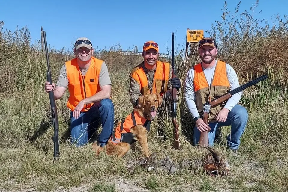 Three hunters and a dog pose with harvested pheasants and chukar in a grassy field.