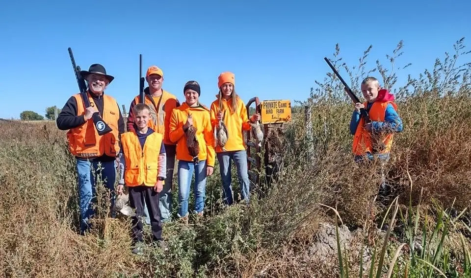 A group of hunters, including adults and children, pose with harvested ring-necked pheasants in a field.