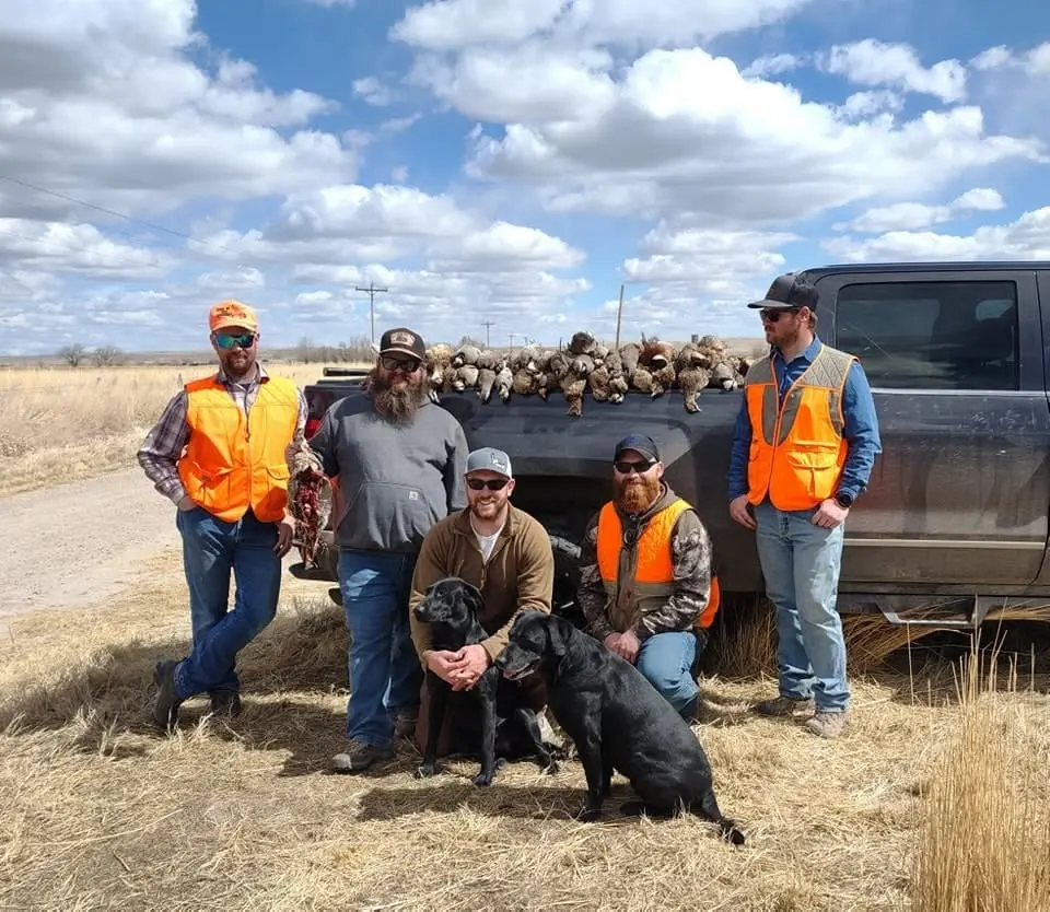 Five hunters and two dogs pose with a truck bed full of harvested pheasants and chukar.
