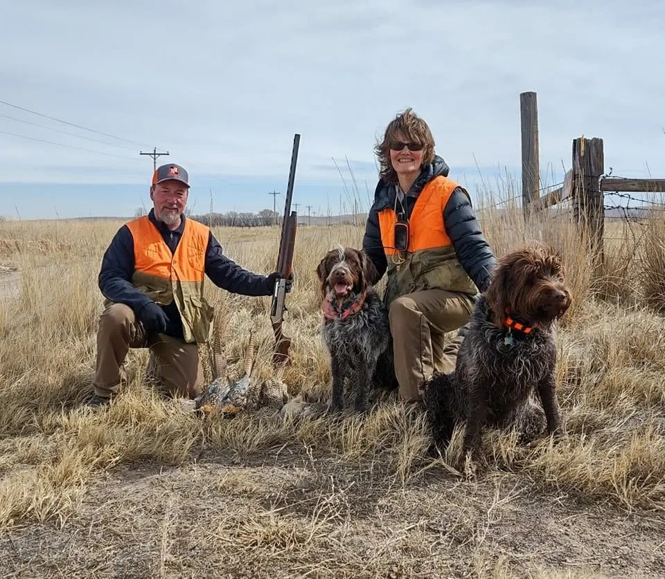 Two hunters and two dogs pose with harvested ring-necked pheasants in a field.