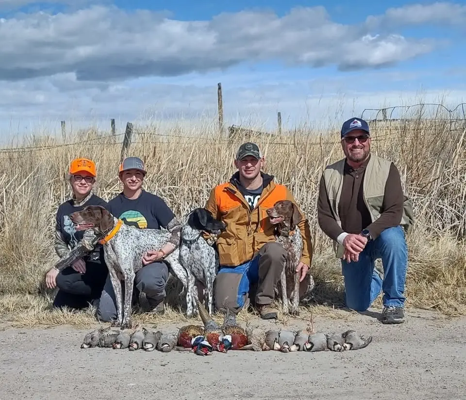 Four hunters and three dogs pose with harvested ring-necked pheasants and chukar partridges in a field.