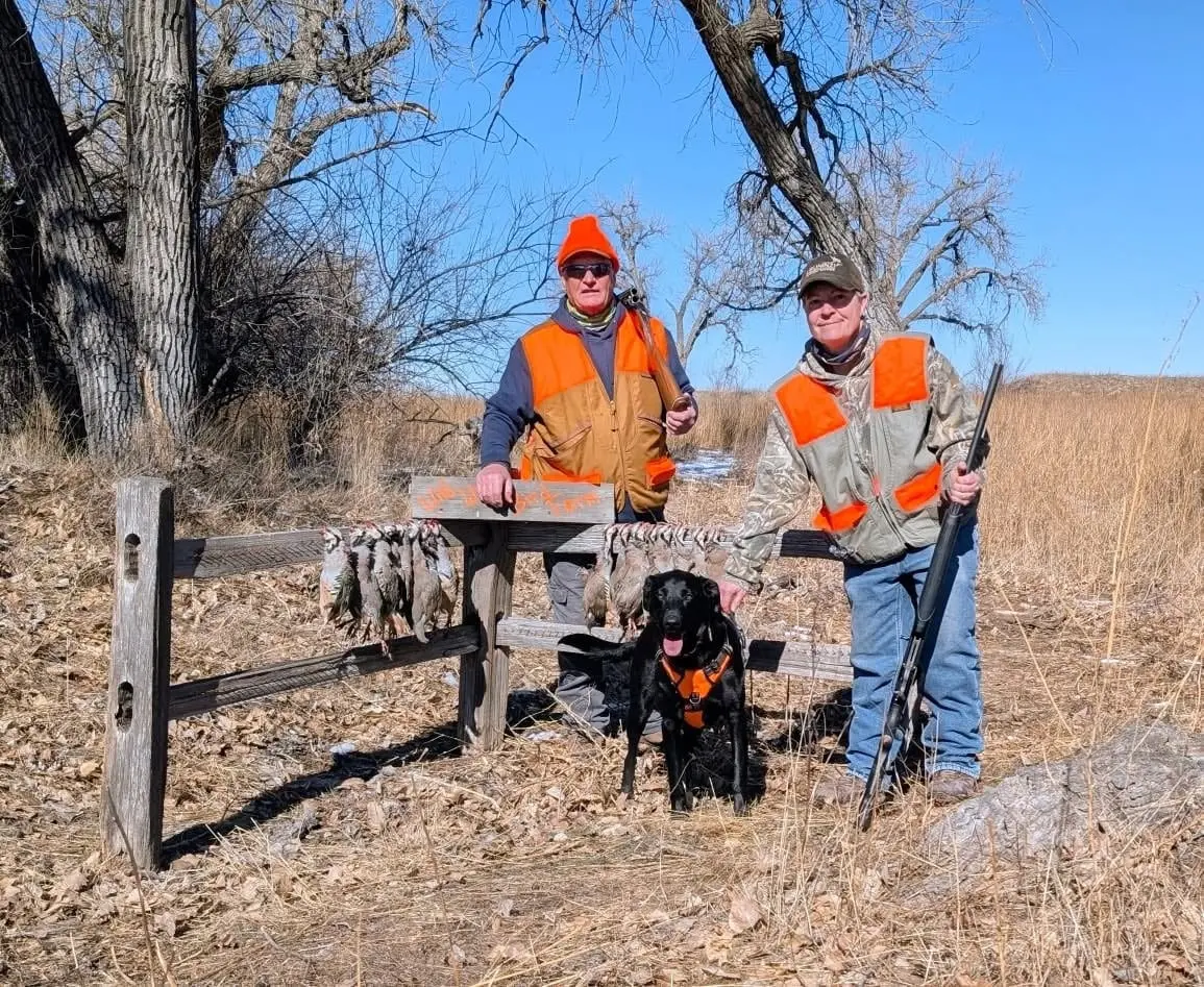 Two hunters and a black dog pose with harvested chukar hanging on a wooden fence in a field.