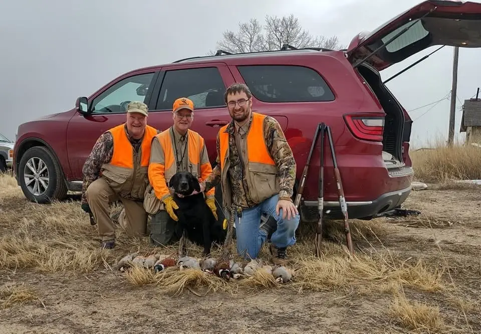 Three hunters and a black dog pose with harvested ring-necked pheasants and chukar partridges in front of a red SUV.