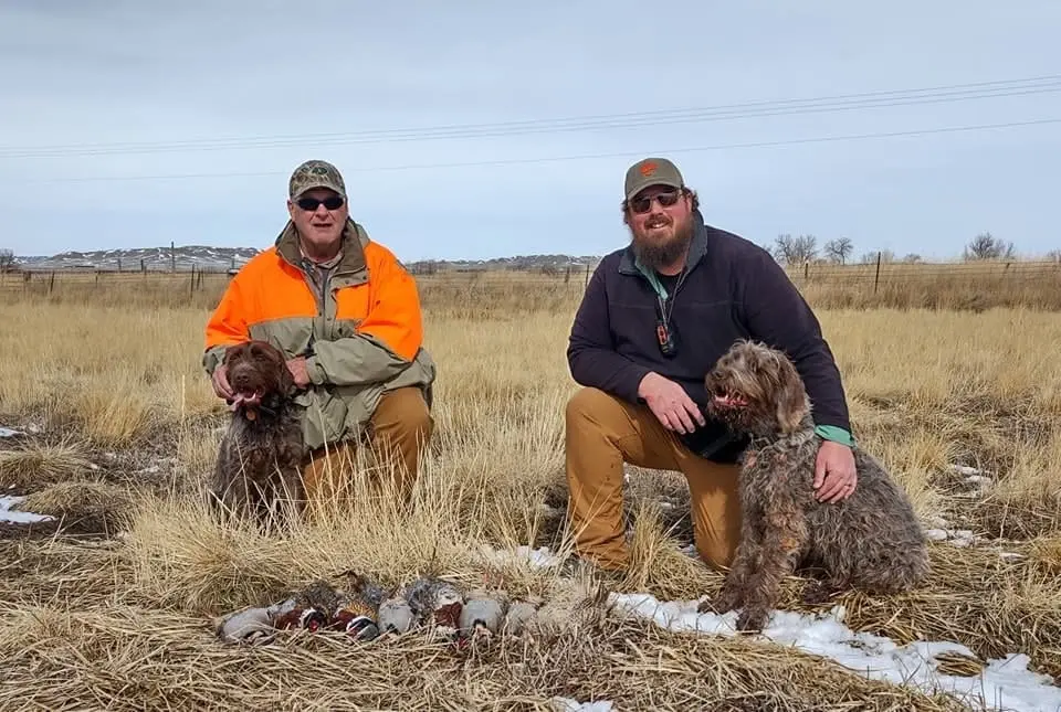 Two men and two dogs pose with harvested pheasants and chukar in a field.
