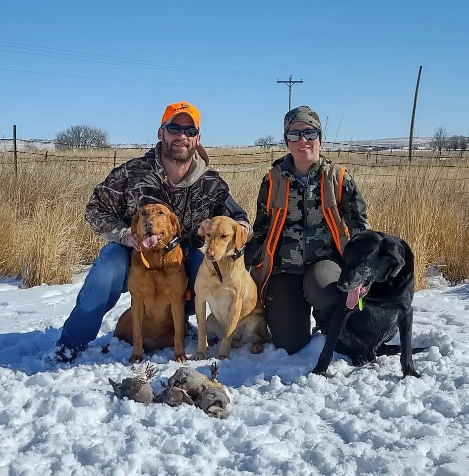 Two hunters pose with four hunting dogs and several harvested chukar partridges in a snowy field.