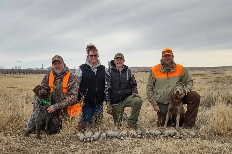 A group of hunters and their dogs pose with harvested chukar partridges in a field.
